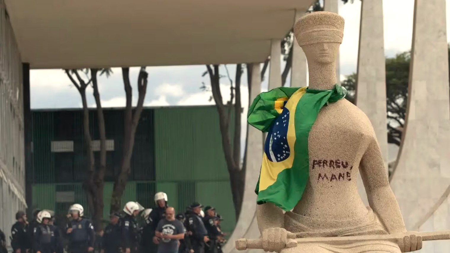 A escultura A Justiça, em frente ao Supremo Tribunal Federal, com uma bandeira do Brasil amarrada no ombro e a frase "perdeu, mané" escrita sobre a pedra. Ao fundo, policiais de capacete.