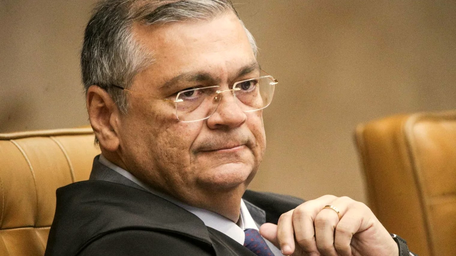 O ministro Flávio Dino sentado na bancada do plenário do Supremo Tribunal Federal, em close, de toga, olhando para o lado com as mãos unidas à frente.