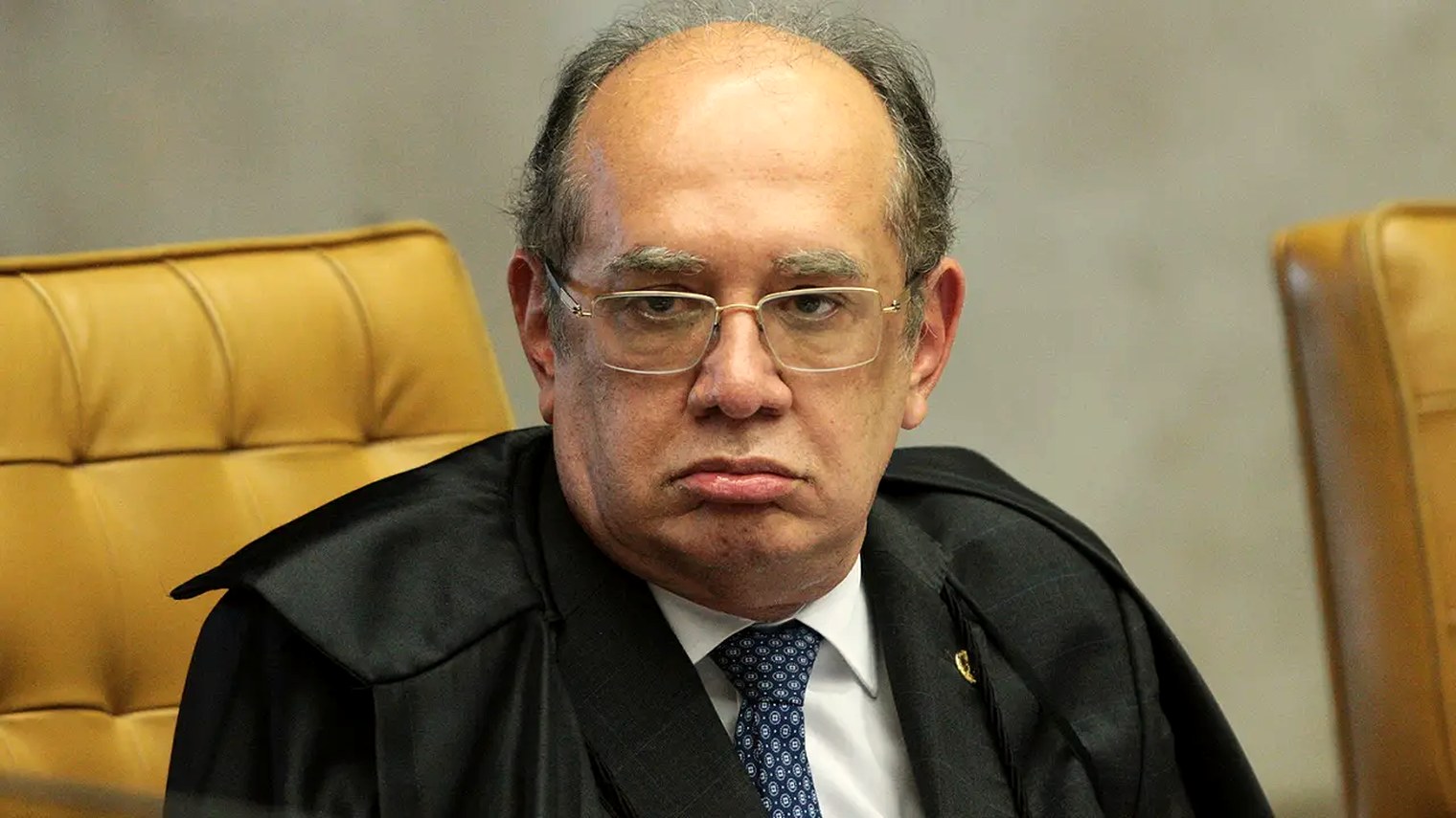 O ministro Gilmar Mendes sentado na bancada do plenário do Supremo Tribunal Federal, em close, de toga, com expressão fechada.